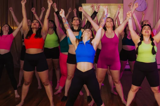A group of women participating in an energetic dance workout. They are standing with their arms raised in the air, wearing colorful workout outfits, and engaging enthusiastically in the activity.