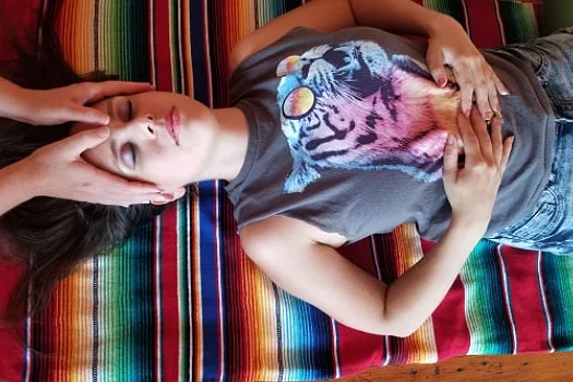 A serene woman receiving a Reiki treatment at Hustle and Flow studio. She is lying down on a colorful striped blanket with her eyes closed, wearing a tank top with a tiger graphic and denim jeans. The practitioner's hands are gently placed on her head.