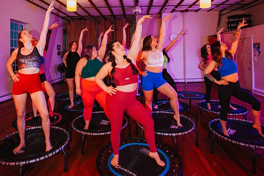A group of women participating in a trampoline workout class at Hustle and Flow studio. They are standing on mini trampolines, wearing colorful workout outfits, and striking a pose with one arm raised.