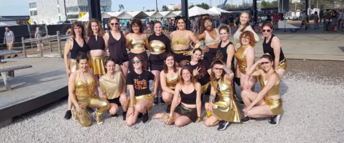 A group of dancers from Hustle and Flow studio in Portland, Maine, posing together outdoors. They are wearing coordinated black and gold outfits, smiling.